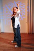 A young woman dances in a studio, her white shirt flowing as she moves, a geometric pattern projected on the wall behind her photo