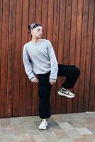 A young man dances in front of a wood panel wall, wearing a grey sweater, black pants, and white sneakers photo