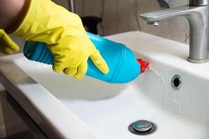 The hands of a girl in a yellow cap with a blue bottle of detergent clean and wash the sink from limescale and dirt. Cleaning company services, close-up photo