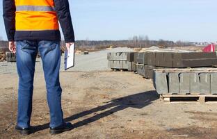An engineer with a folder in the signal vest checks the quality of the curb for the construction of a new road, inspector photo
