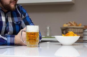 A man with a beard is sitting in the kitchen, on the table is a glass of beer and chips, junk food, domestic photo