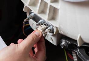 The hand of the master disconnects the electrical chip from the temperature sensor of the washing machine. Replacing the thermostat, close-up, industry photo