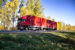 The new red truck with a semi-trailer transports a lot of bulk cargo. Cargo transportation on a country road against the backdrop of a forest in autumn. Copy space for text photo