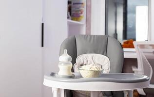 A baby bottle with milk and a plate with dry milk formula enriched with bifidobacteria, vitamins and minerals stands on a gray high chair in the children's room. Copy space for text photo