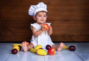 baby girl in a chef's cap is sitting on the floor, holding an apple and smiling. apples, mangoes, bananas, plums lie on the floor photo