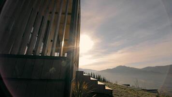Wooden structure with sunlight and mountain view at sunrise - A modern wooden structure with sunlight streaming through slats, set against a backdrop of rolling hills and morning light. video