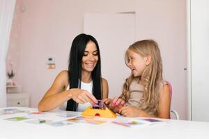 Smiling mother and a little daughter play a logical strategy board game together at the table in the nursery, a happy family is engaged in logical activities at home photo