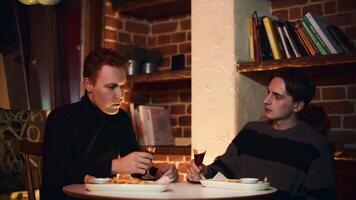 Two friends are sitting in restaurant, chatting and drinking alcoholic drink. Media. Two young men are drinking alcoholic tincture in restaurant. Cozy meeting of two friends in restaurant with video