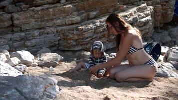 Young mother and a little baby boy sitting on the beach and playing with sand. Creating. Family vacation with children. video