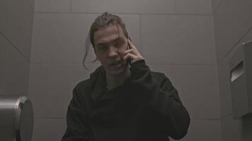 Young man sits on toilet and talks on phone. Stock. Man in black walks into toilet stall and talks on phone. Young man is talking aggressively on phone in toilet video