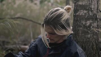 Young man sits tired by tree in forest. Stock. Young man is lost in forest and sits in despair under tree. Man is sitting by tree with desperate look and loss video