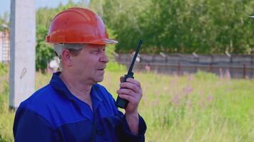 Portrait of mature construction superintendent using transmitter for talking to workers. Clip. Man in protective uniform and helmet outdoors. video
