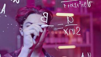 Young man solves mathematical equations on blackboard. Media. Young man solves complex equations on transparent board. Genius writes equations on blackboard in neon light video