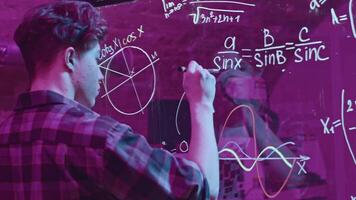 Young man solves mathematical equations on blackboard. Media. Young man solves complex equations on transparent board. Genius writes equations on blackboard in neon light video