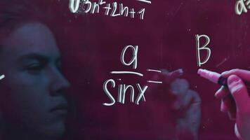 Young man solves equations. Media. Close-up of young man's reflection on transparent writing board with marker. Man solves mathematical equations with marker on blackboard video