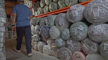 Young man passing by a stack of rolls of fabric lies in the warehouse of a garment factory. Media. Garment factory for the production of warm clothes. video