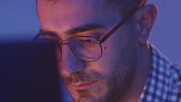 Close-up of young tired man working in front of computer. Media. Young man with glasses is programming at computer. Tired programmer is working at computer video