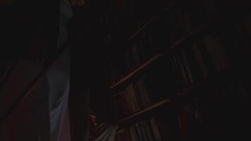Low angle view of of a couple slowly walking in a dark library. Stock footage. Man and woman passing by shelves with books. video