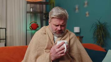 Sick unhealthy man wrapped in plaid sit alone shivering from cold on sofa drinking hot tea at home video