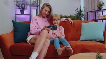 Child kid daughter having fun with mother or nanny enjoying using smartphone face mask app at home video