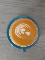 a cup of latte placed on a blue saucer, sitting on a wooden table. The latte has a delicate latte art design on its frothy surface, resembling a stylized flower or leaf. photo