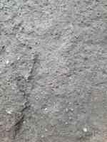 a close-up of a rough, textured surface, likely a rock or a concrete slab. It appears weathered and worn, with various cracks, crevices, and small pebbles embedded within it. photo