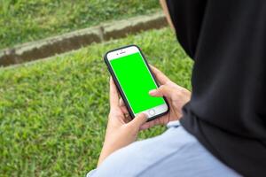 A joyful woman in a black hijab and blue shirt smiles brightly while holding a smartphone, standing at the edge of a beautiful park sidewalk photo