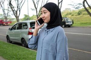 A woman wearing a black hijab and blue shirt smiles while on a call, occasionally looking at the camera. She appears cheerful and engaged, exuding a positive, friendly vibe photo