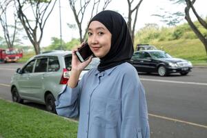 A woman wearing a black hijab and blue shirt smiles while on a call, occasionally looking at the camera. She appears cheerful and engaged, exuding a positive, friendly vibe photo