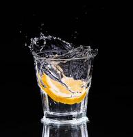 Slice of lemon splashing into a glass of water with a spray of water droplets in motion suspended in the air above the glass on a dark background. photo