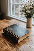A simple, closed book resting on a rustic wooden stool in a sunlit room with a window photo