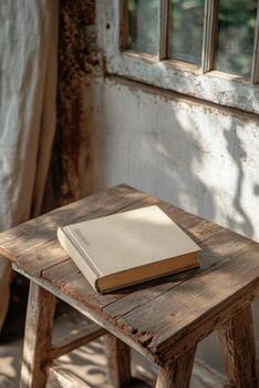 A simple, closed book resting on a rustic wooden stool in a sunlit room with a window photo