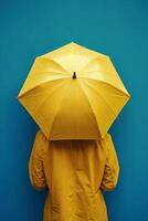 A person in a yellow raincoat stands under a bright yellow umbrella against blue photo