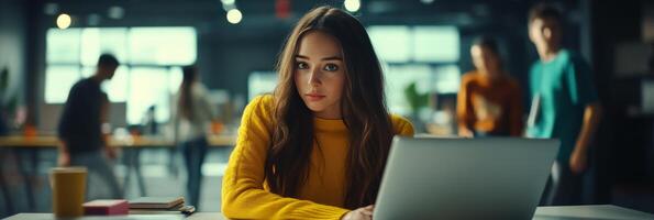 Young woman focused on laptop in modern workspace during daytime with colleagues in background photo