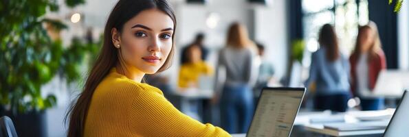 Young woman focused on laptop in modern workspace during daytime with colleagues in background photo