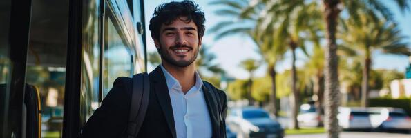 Smiling young man wearing suit stands near bus under palm trees during sunny day photo