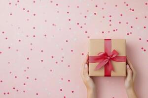 A person holds a beautifully wrapped gift with a red bow over a confetti filled pink background photo