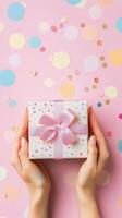 A person holding a blue gift box with pink ribbon on a pastel pink table covered in confetti photo