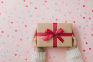 A person holds a beautifully wrapped gift with a red bow over a confetti filled pink background photo