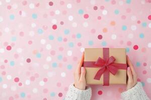 A person holds a beautifully wrapped gift with a red bow over a confetti filled pink background photo