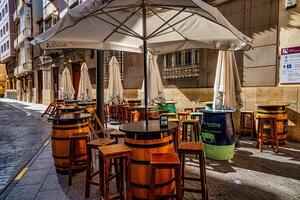 Outdoor cafe tables and chairs are set up on a quaint cobblestone street, featuring barrels as table bases and shade umbrellas overhead. photo