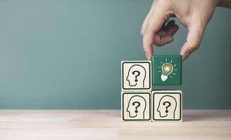 A hand picks up light bulb icon inside a wooden block placed around a human icon with question. Concept of seeking knowledge, intelligent problem solving, good ideas, and new innovation. photo
