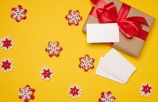 Christmas gift wrapped in red ribbon on yellow background, stack of white paper business cards. Top view photo