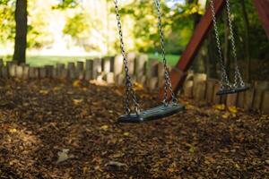 Empty black swing on children playground in the park in autumn season. Missing child. Lonely swing in sunny autumn day. Golden foliage in late fall. Beautiful nature. photo