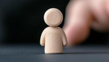 A wooden figure stands on a dark surface as a finger reaches out towards it photo