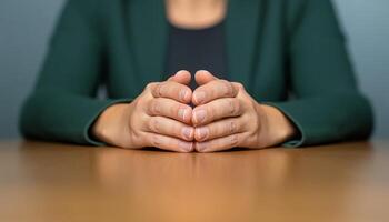 A person sits at a wooden table with hands clasped together in a thoughtful manner photo