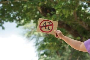 Close up hand holds paper card with No AI, prohibition sign, outdoor nature background. Concept, disagree, anti or don't support to use AI or Artificial Intelligence in daily life. photo