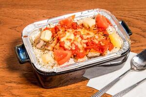 Spanish baked Pork Chop Rice topped with melted cheese, pineapple chunks, and tomato sauce served in foil container dish isolated on wooden table side view of spanish food photo