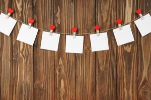 Clothespins with red hearts and empty sheets of paper on string against brown wooden background photo
