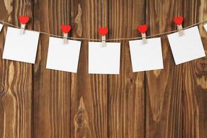 Clothespins with red hearts and empty sheets of paper on string against brown wooden background photo